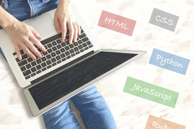 ノートパソコンでタイピングする手元と、周囲に配置されたHTML、CSS、Python、JavaScript、Rubyなどのプログラミング言語名カード